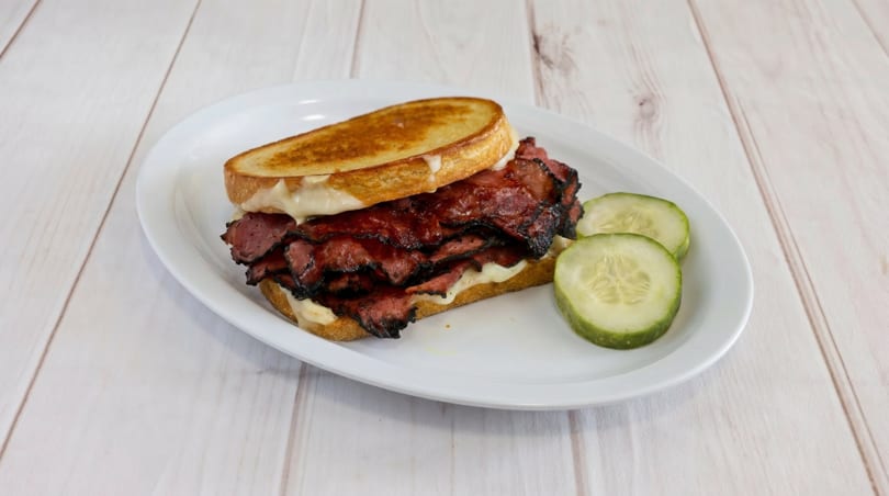 A grilled pastrami sandwich with melted cheese and two cucumber slices on a white plate.