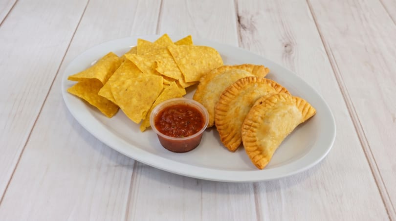 A white platter with golden empanadas, tortilla chips, and a cup of salsa on a wooden table.