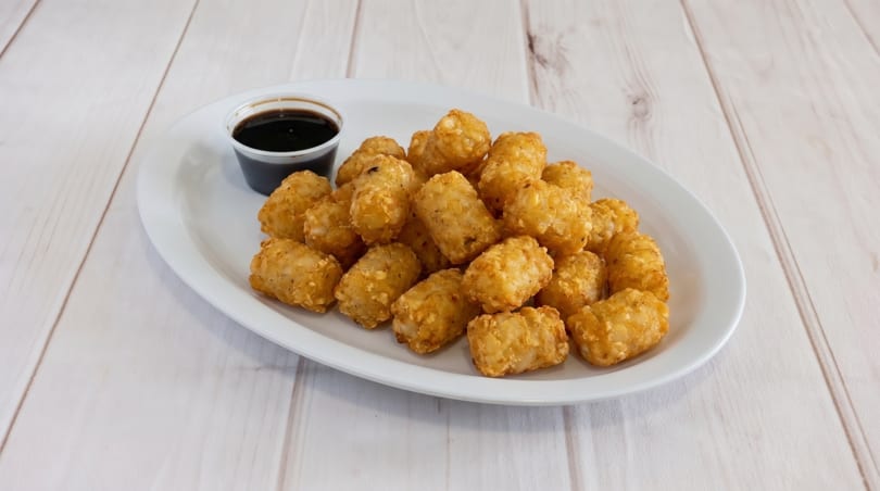 A white oval plate filled with golden-brown tater tots and a small cup of dark dipping sauce.