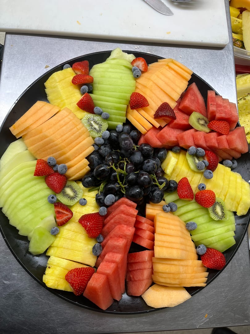 A large black platter filled with an assortment of sliced fresh fruits including melon, pineapple, and berries.
