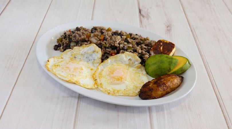 Gallo pinto with two fried eggs, fried plantains, avocado slices, and fried cheese on a white plate.