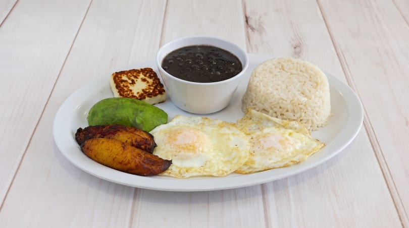A white plate with fried eggs, rice, black beans, avocado, fried plantains, and grilled cheese.