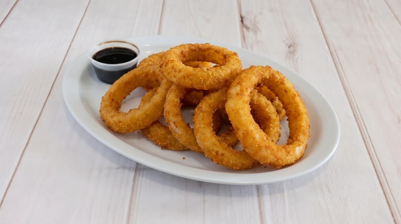 Golden-brown onion rings piled on a white plate with a side of dark dipping sauce.